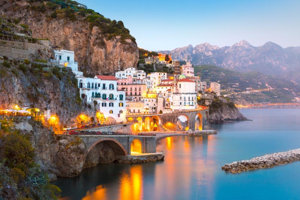 The town of Amalfi seen from the water at dusk, pastel buildings stacked against steep cliffs above the harbour.